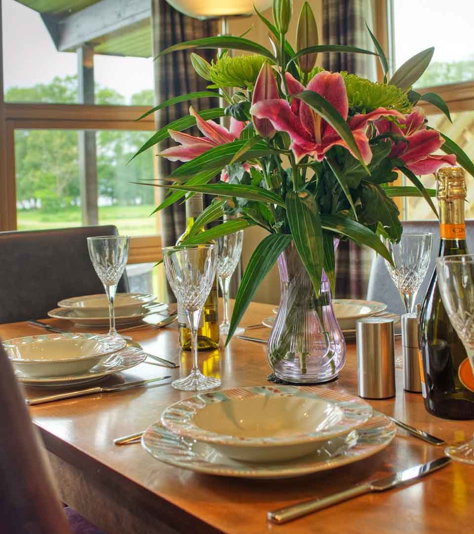 A table set for dining with a vase of lilies and a view out to Loch Lomond in a chalet at Loch Lomond Waterfront