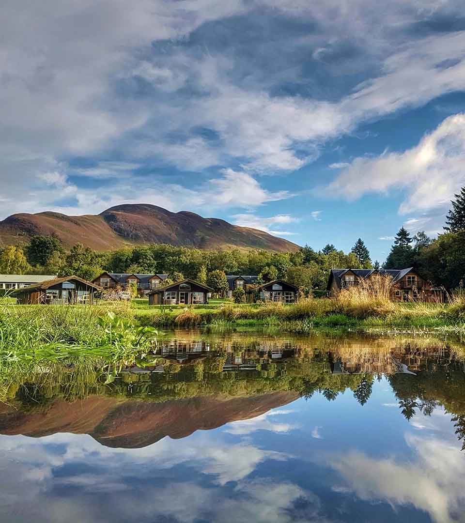 A view over a glass still pond of Loch Lomond Waterfront with Conic Hill in the background